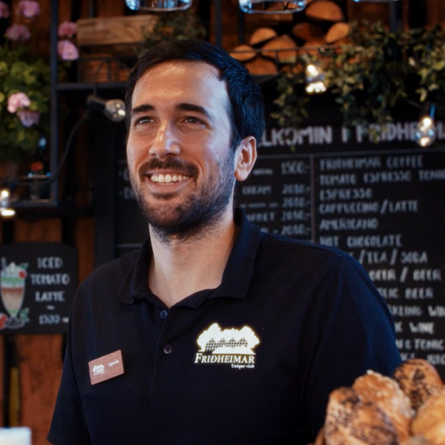 Smiling man in a black Fridheimar shirt standing in a cafe with breads and a menu board in the background.
