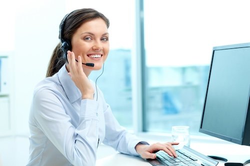 Smiling woman wearing headset working at a computer in a bright office.
