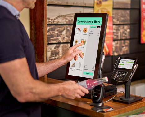 Person using self-checkout touchscreen at a convenience store while holding a bottled beverage.