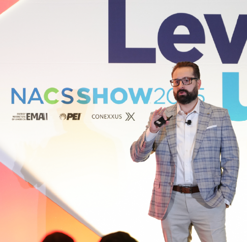 Man in a plaid blazer and glasses giving a presentation on stage with NACS SHOW 2025 logo in the background.
