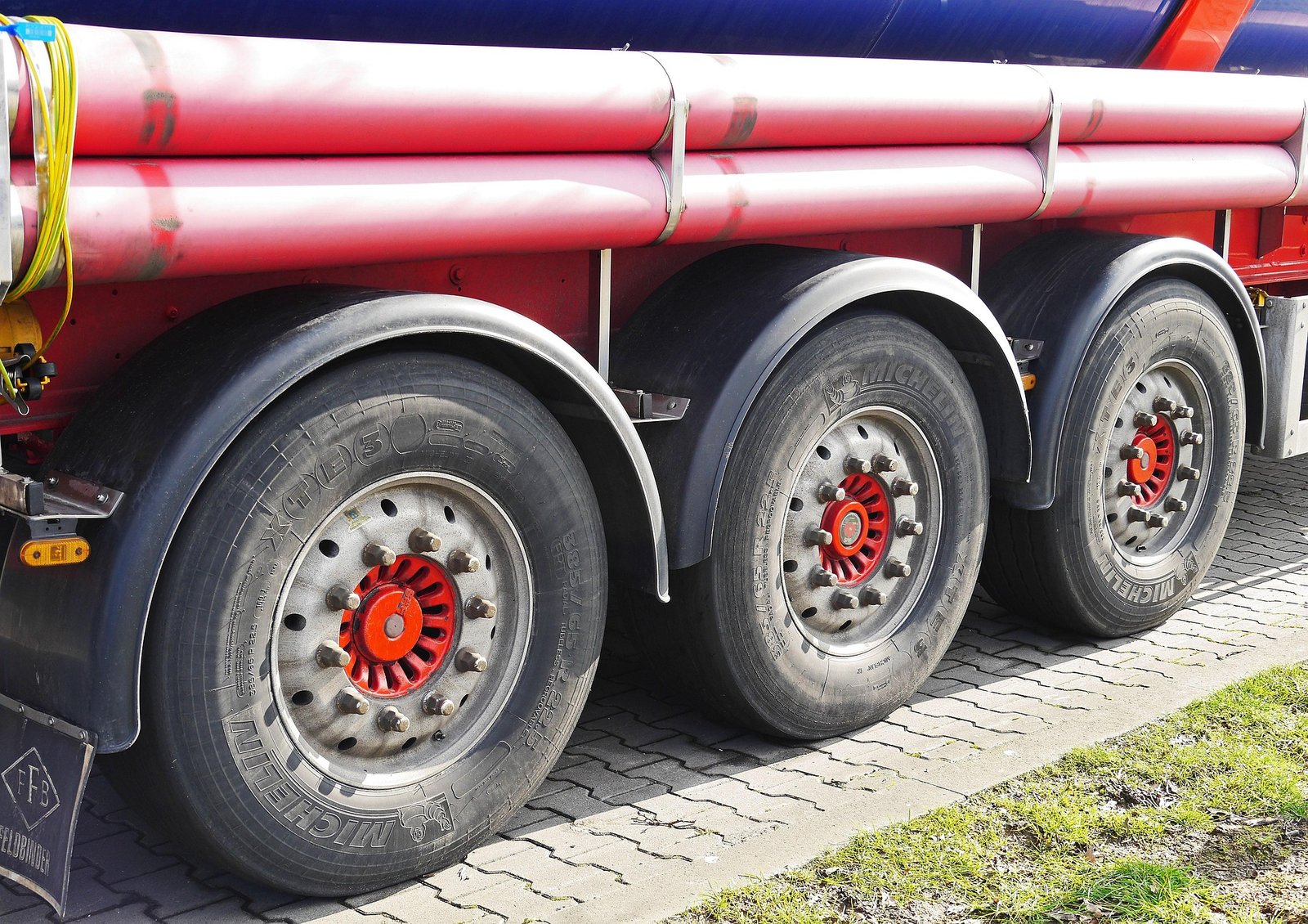 Close-up of three large Michelin tires with red hubs on a red tanker truck parked on a paved surface with grass beside it.