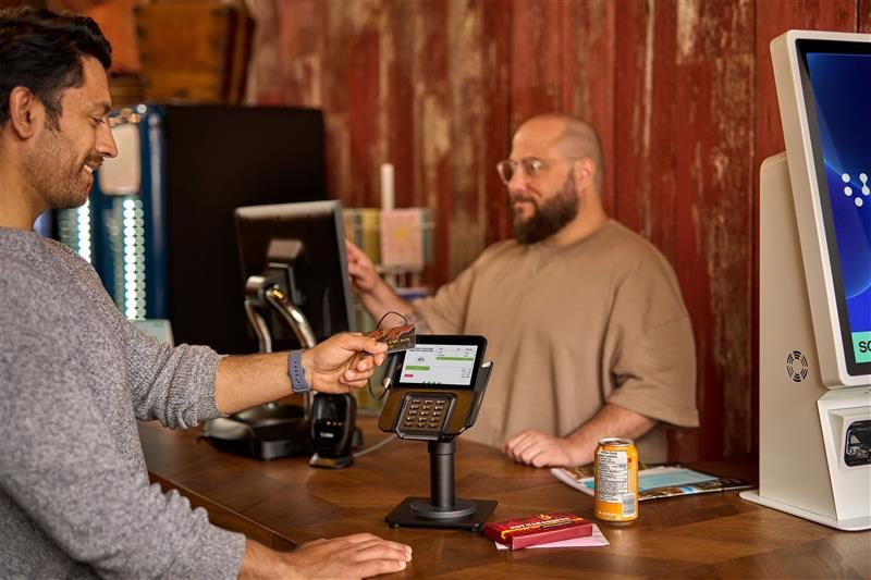 Smiling customer holding a card near a card payment terminal on a wooden counter with a cashier and self-service kiosk in the background.
