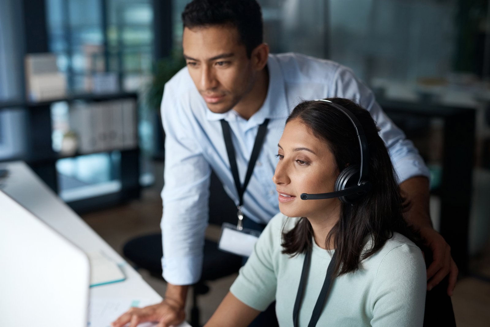 Woman wearing a headset working at a computer with a man standing beside her, both focused on the screen in a modern office.