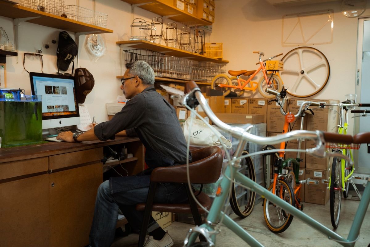 Man sitting at a desk working on an iMac computer in a bicycle shop with bikes and boxed bike parts in the background.
