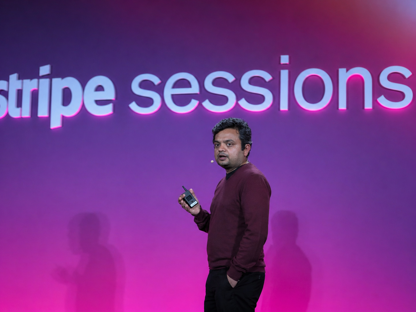 Man holding a wireless microphone speaking on stage with 'stripe sessions' text illuminated in pink behind him.